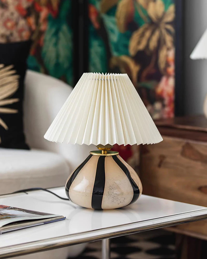 Pleated Cavolo table lamp with a striped ceramic base and cream pleated shade, styled on a white coffee table in an eclectic living room with floral decor and colorful accents.