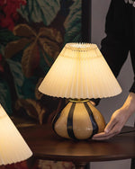 Load image into Gallery viewer, Pleated Cavolo table lamp with a glowing pleated shade and striped ceramic base, placed on a round wooden table as a person adjusts its position, with floral decor in the background.
