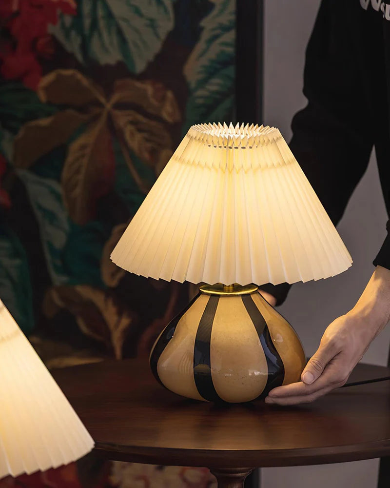 Pleated Cavolo table lamp with a glowing pleated shade and striped ceramic base, placed on a round wooden table as a person adjusts its position, with floral decor in the background.