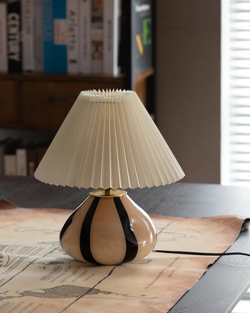 Pleated Cavolo table lamp with a striped ceramic base and cream pleated shade, placed on a desk over a vintage map, creating a cozy study space with shelves of books in the background.