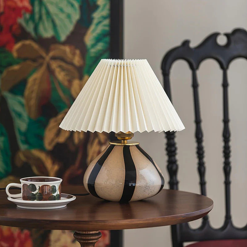 Pleated Cavolo table lamp with a striped ceramic base and cream pleated shade, placed on a round wooden table alongside a decorative teacup, with a black vintage chair and floral backdrop adding charm to the scene.