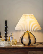 Load image into Gallery viewer, Pleated Cavolo table lamp with a glowing pleated shade and striped ceramic base, placed on a wooden vanity table styled with perfume, a decorative tray, a pearl necklace, and a sleek black candleholder.
