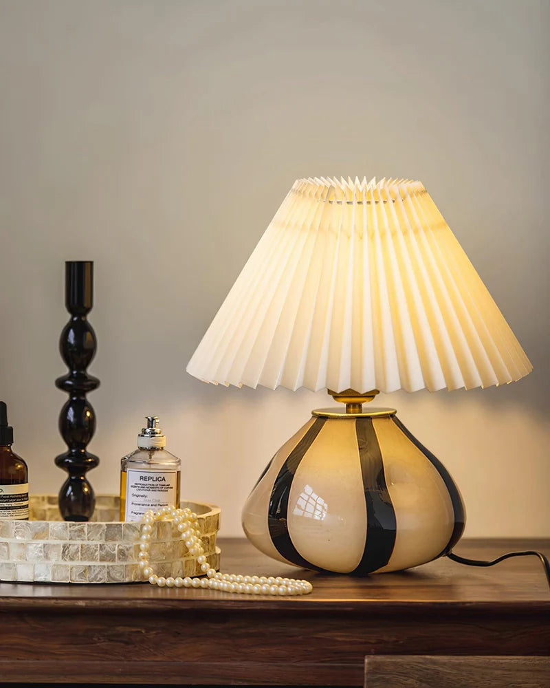 Pleated Cavolo table lamp with a glowing pleated shade and striped ceramic base, placed on a wooden vanity table styled with perfume, a decorative tray, a pearl necklace, and a sleek black candleholder.