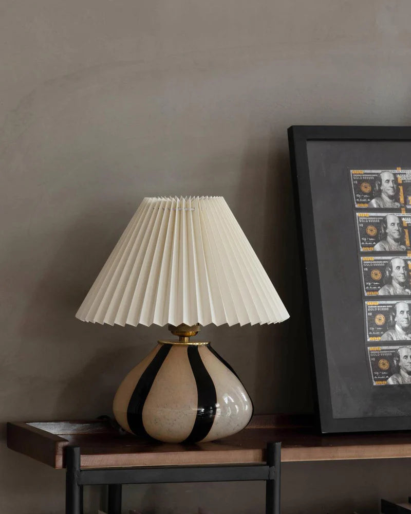 Pleated Cavolo table lamp featuring a ceramic base with bold black stripes and a cream pleated lampshade, styled on a wooden shelf next to framed wall art.