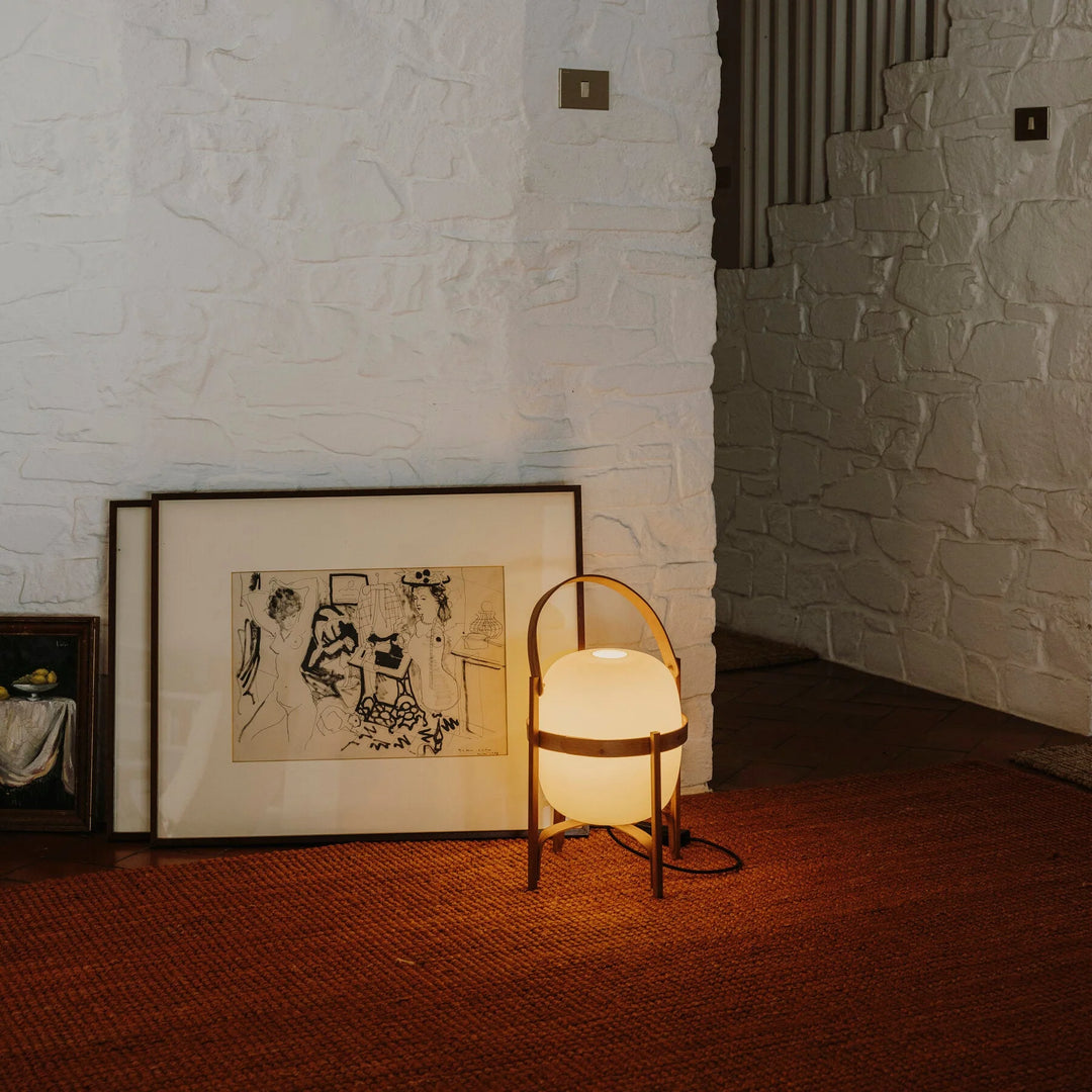 Portable lantern-style glass lamp with a soft glow, featuring a wooden frame and leather handle, placed on a textured rug next to framed artwork resting against a white brick wall, adding a warm ambiance to the display.