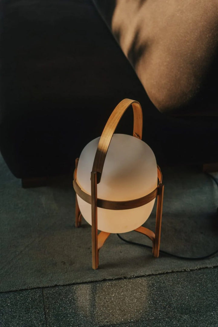 Portable lantern-style glass lamp with a soft glow, featuring a wooden frame and leather handle, placed on a rug next to a dark sofa, creating a warm and inviting ambiance in a cozy corner.