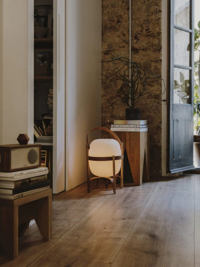 Portable lantern-style glass lamp with a warm glow, featuring a wooden frame and leather handle, placed on a wooden floor beside a rustic side table with books and a potted plant, creating a cozy and inviting living room ambiance.