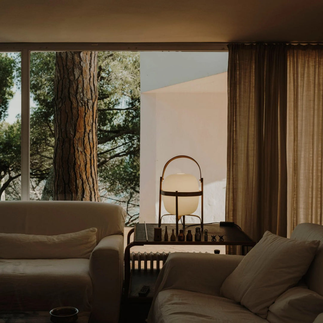 Portable lantern-style glass lamp with a soft, warm glow, featuring a wooden frame and leather handle, placed on a side table between white sofas in a serene lounge area with large windows overlooking a forest view.