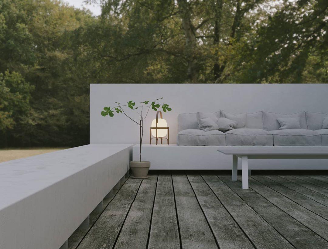 Portable lantern-style glass lamp with a warm glow, featuring a wooden frame and leather handle, placed on an outdoor seating area with a minimalist white sofa, a potted plant, and surrounded by lush greenery.