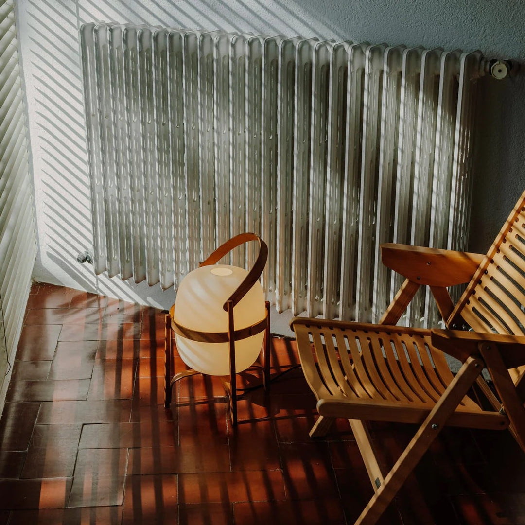 Portable lantern-style glass lamp with a warm glow, featuring a wooden frame and leather handle, placed on a sunlit floor near a folding wooden chair and a vintage radiator, creating a cozy, natural ambiance.