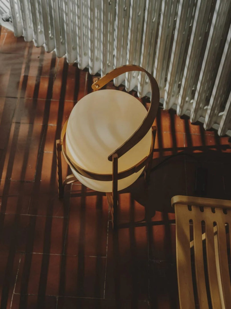 Top-down view of a portable lantern-style glass lamp with a warm glow, featuring a wooden frame and leather handle, placed on a terracotta-tiled floor with striped sunlight shadows creating a cozy atmosphere.