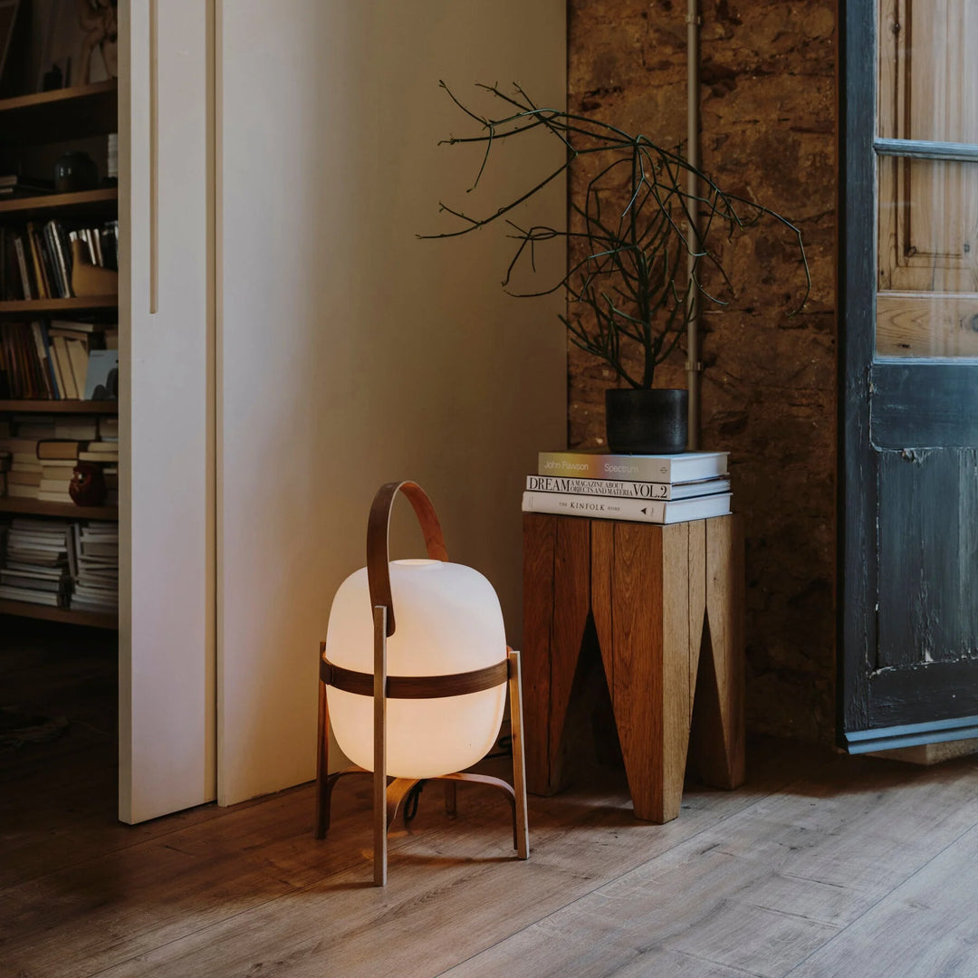 Portable lantern-style glass table lamp with a warm glow, featuring a wooden frame and leather handle, placed on the floor beside a rustic wooden side table decorated with books and a potted plant.