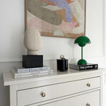 Load image into Gallery viewer, A minimalist setup featuring a green rechargeable LED desk lamp, a small black candle holder, and a beige sculptural piece. The lamp is placed on a stack of stylish books, beside a painting, on a white dresser.
