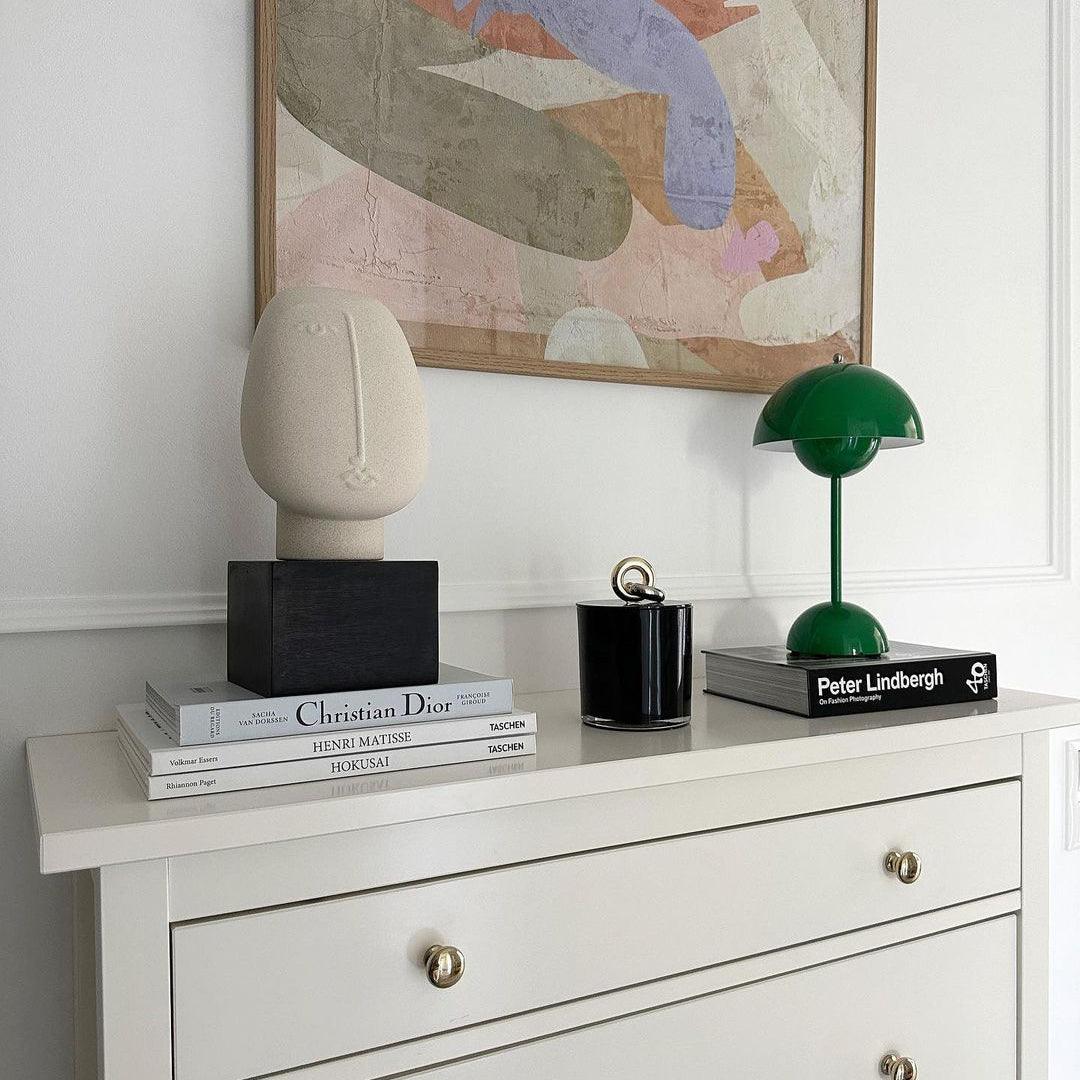 A minimalist setup featuring a green rechargeable LED desk lamp, a small black candle holder, and a beige sculptural piece. The lamp is placed on a stack of stylish books, beside a painting, on a white dresser.