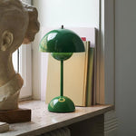 Load image into Gallery viewer, A vibrant green rechargeable desk lamp with a dome-shaped shade and a spherical base. The lamp is placed on a stone shelf near a sculpture, books, and a window, contributing to a minimalist and stylish setting. The lamp is a perfect blend of functionality and modern design.
