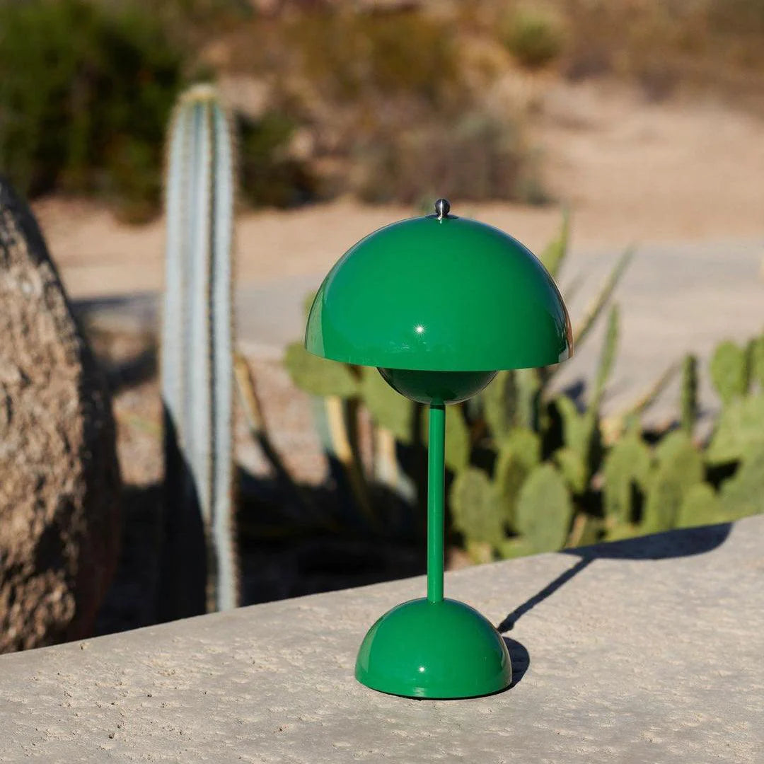 A small, vibrant green rechargeable desk lamp with a rounded dome-shaped shade, standing on a spherical base. It features a minimalistic design, suitable for modern decor. The lamp is positioned outdoors on a stone surface with cacti in the background.