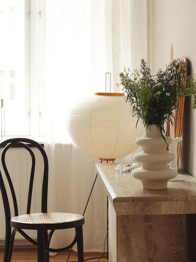 A cozy interior featuring a rice paper floor lamp on tripod legs beside a marble console table with a sculptural white vase filled with fresh greenery, a black cane chair, and soft natural light streaming through sheer curtains.