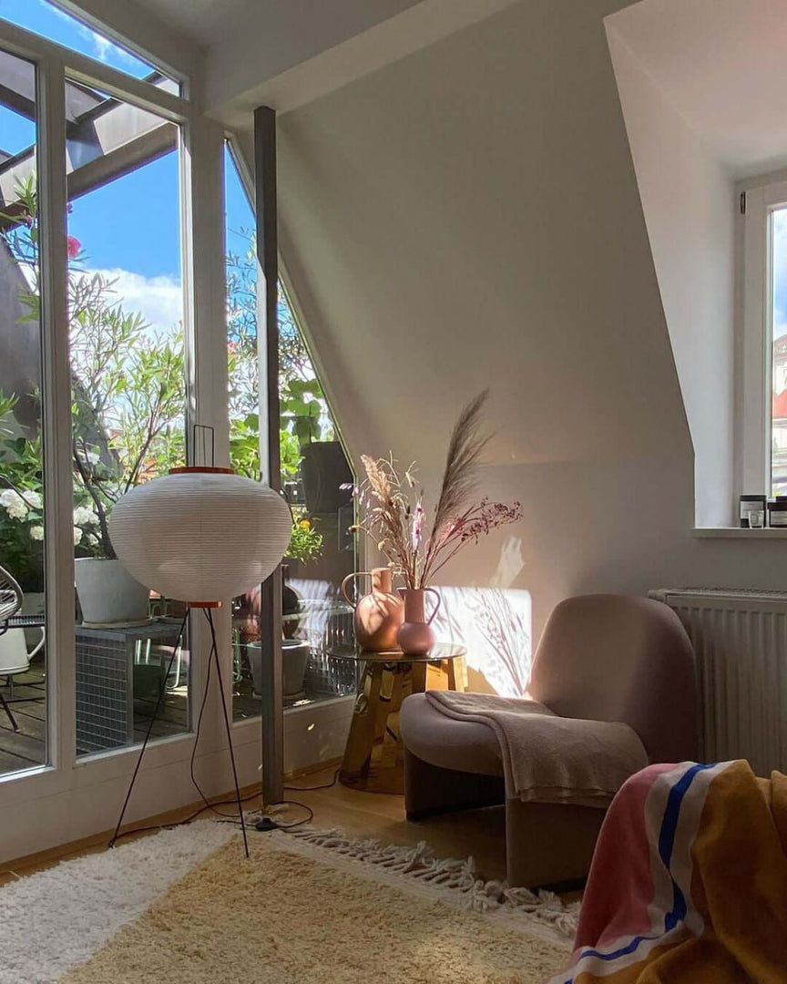 A cozy sunlit nook featuring a rice paper floor lamp, a plush armchair with a blanket, decorative vases with pampas grass on a gold side table, and natural light streaming through glass doors leading to a terrace.