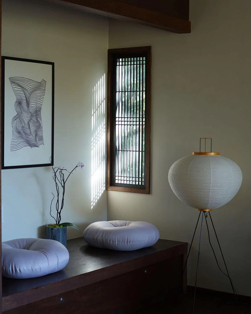 A minimalist interior corner with a rice paper floor lamp on tripod legs, a low wooden bench adorned with round white cushions, a modern vase holding a branch, and natural light streaming through a wooden slatted window.