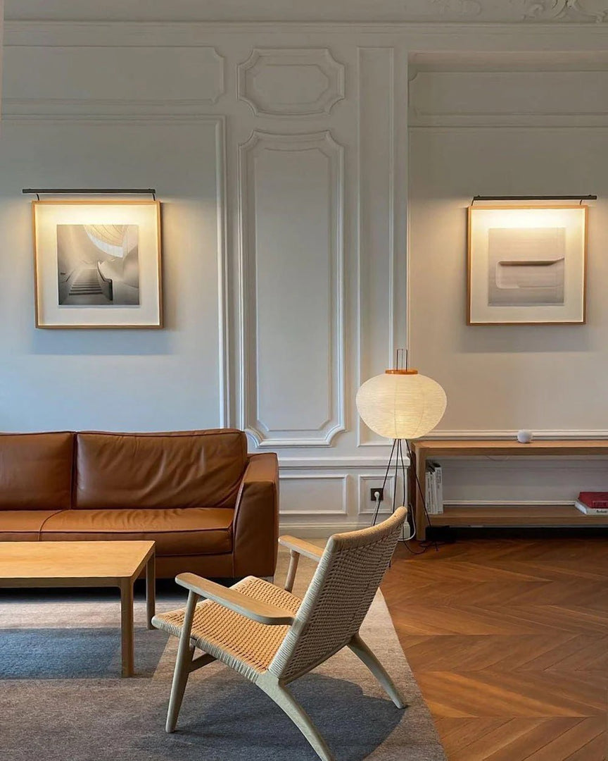 A modern living room with a rice paper floor lamp on tripod legs, a brown leather sofa, a woven lounge chair, a wooden coffee table, and ornate white wall paneling complemented by framed art and warm lighting.