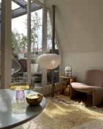 Load image into Gallery viewer, A sunlit room with a rice paper floor lamp on tripod legs near a glass door leading to a terrace, featuring a plush armchair, gold and glass decorative items on a marble table, and a soft shaggy rug.
