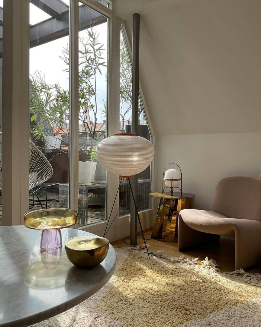 A sunlit room with a rice paper floor lamp on tripod legs near a glass door leading to a terrace, featuring a plush armchair, gold and glass decorative items on a marble table, and a soft shaggy rug.