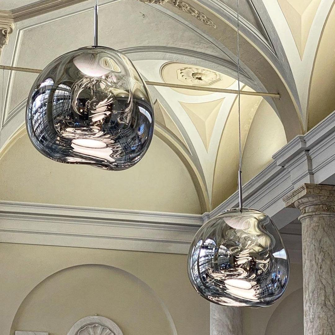 A classical interior featuring two rock pendant lamps with reflective silver surfaces hanging from an ornate ceiling with arches and intricate detailing, complemented by tall marble columns.