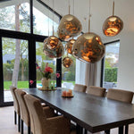 Load image into Gallery viewer, A contemporary dining room featuring an arrangement of rock pendant lamps with reflective gold surfaces hanging above a dark wooden dining table, surrounded by upholstered chairs and decorated with candles and flowers, with large windows overlooking a lush garden.
