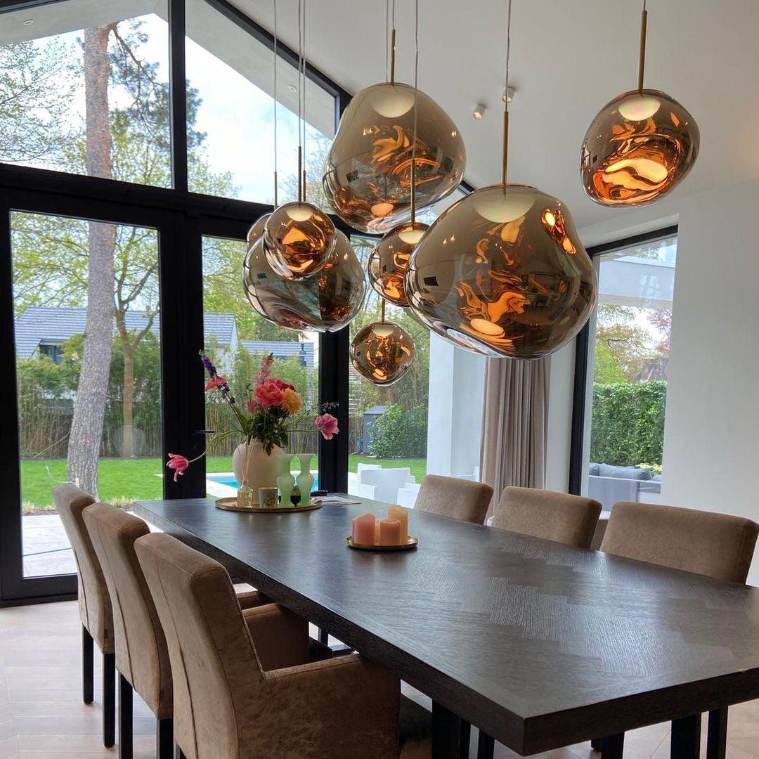 A contemporary dining room featuring an arrangement of rock pendant lamps with reflective gold surfaces hanging above a dark wooden dining table, surrounded by upholstered chairs and decorated with candles and flowers, with large windows overlooking a lush garden.