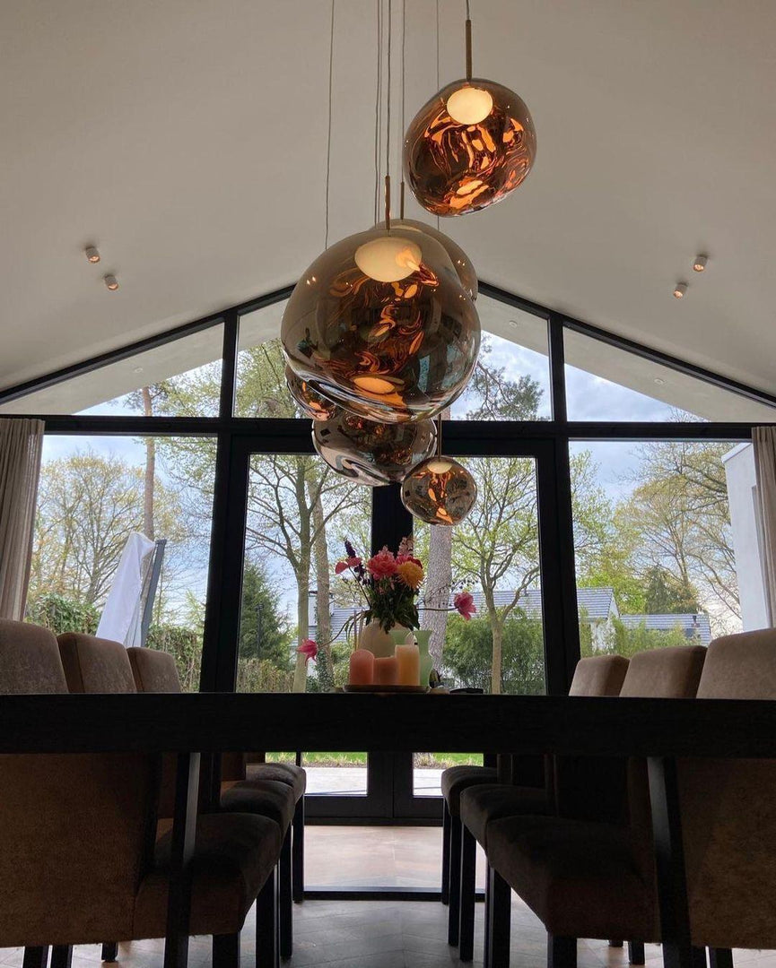 A modern dining room with rock pendant lamps in warm reflective tones hanging above a long table, decorated with flowers and candles, with a backdrop of large glass windows showcasing a garden view.