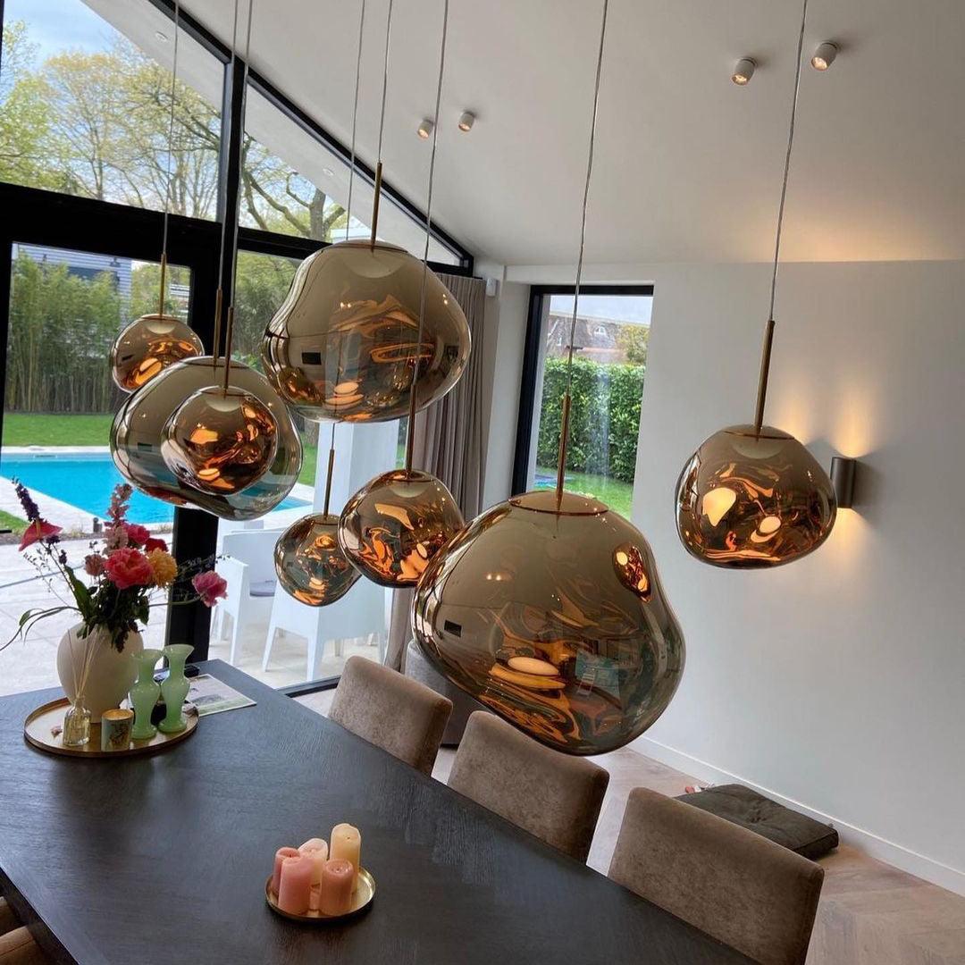 A stylish dining room featuring an arrangement of multiple rock pendant lamps with reflective gold finishes hanging above a dark wooden dining table, decorated with candles and fresh flowers, with large glass doors opening to a pool and garden area.