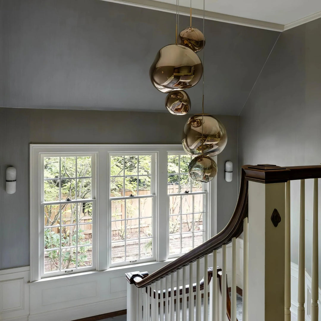 An elegant stairwell featuring a cluster of rock pendant lamps with gold reflective surfaces, hanging in front of large colonial-style windows that offer a view of the garden, complemented by soft gray walls and white wainscoting.