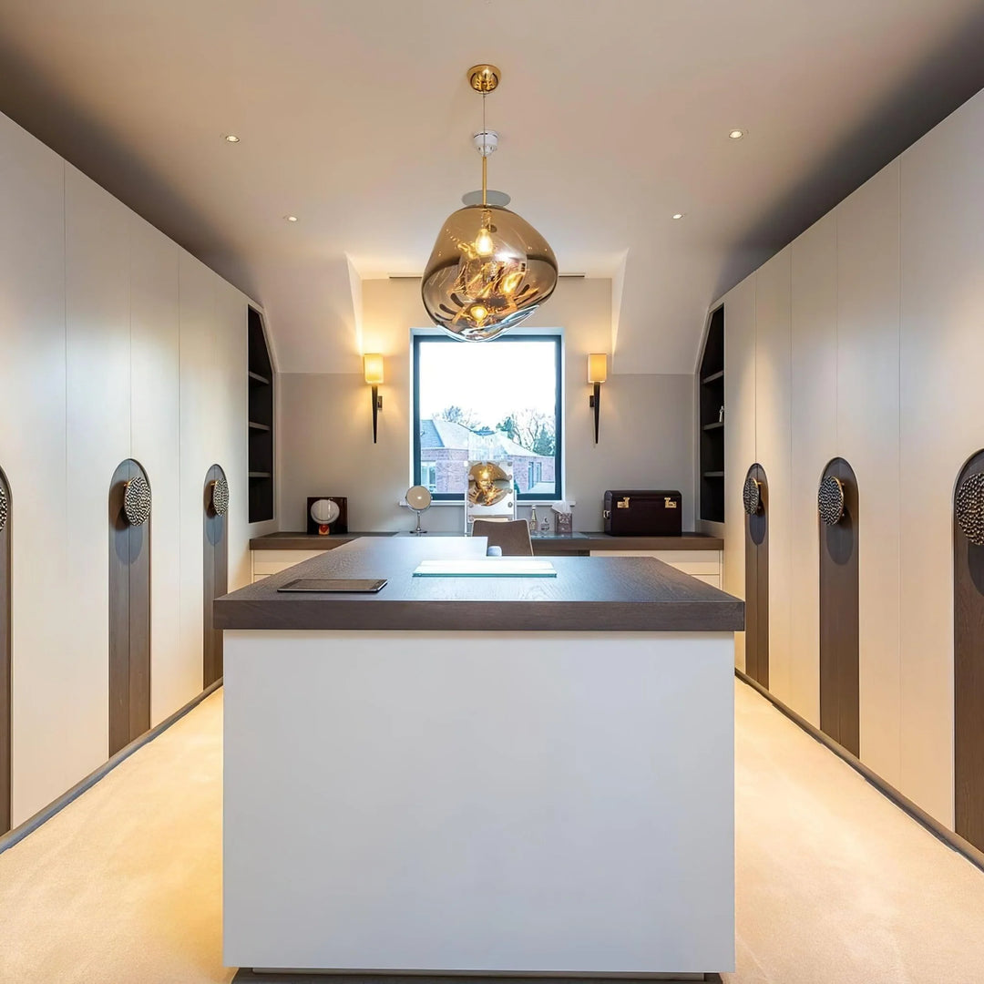 A luxurious dressing room featuring a central island with a reflective rock pendant lamp above, built-in wardrobes with intricate handles, soft recessed lighting, and a large window providing natural light and a view.