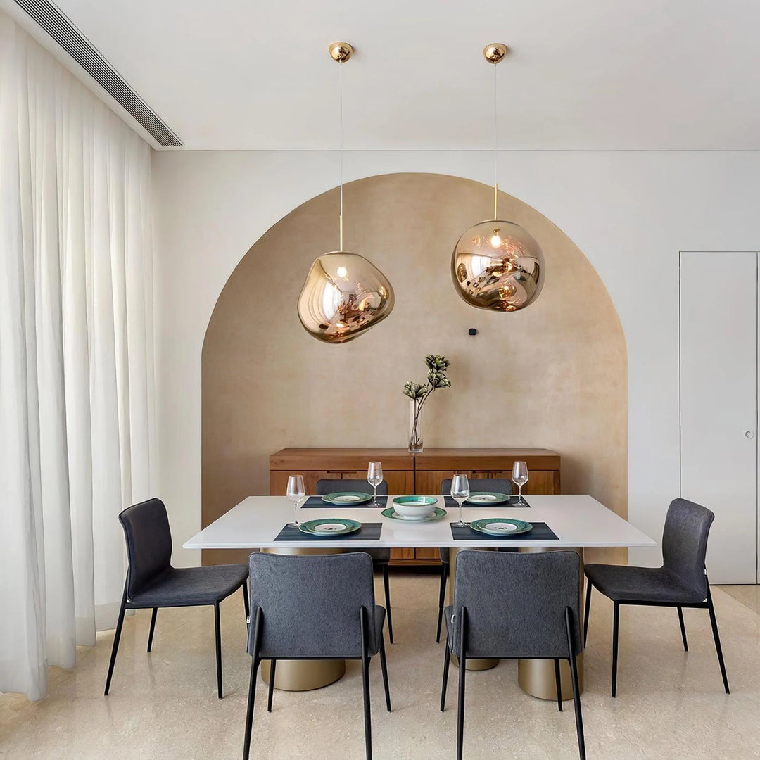 A modern dining area featuring two rock pendant lamps with gold reflective finishes hanging above a white dining table, paired with gray chairs and a minimalist backdrop with an arched wall and a wooden sideboard adorned with a vase.