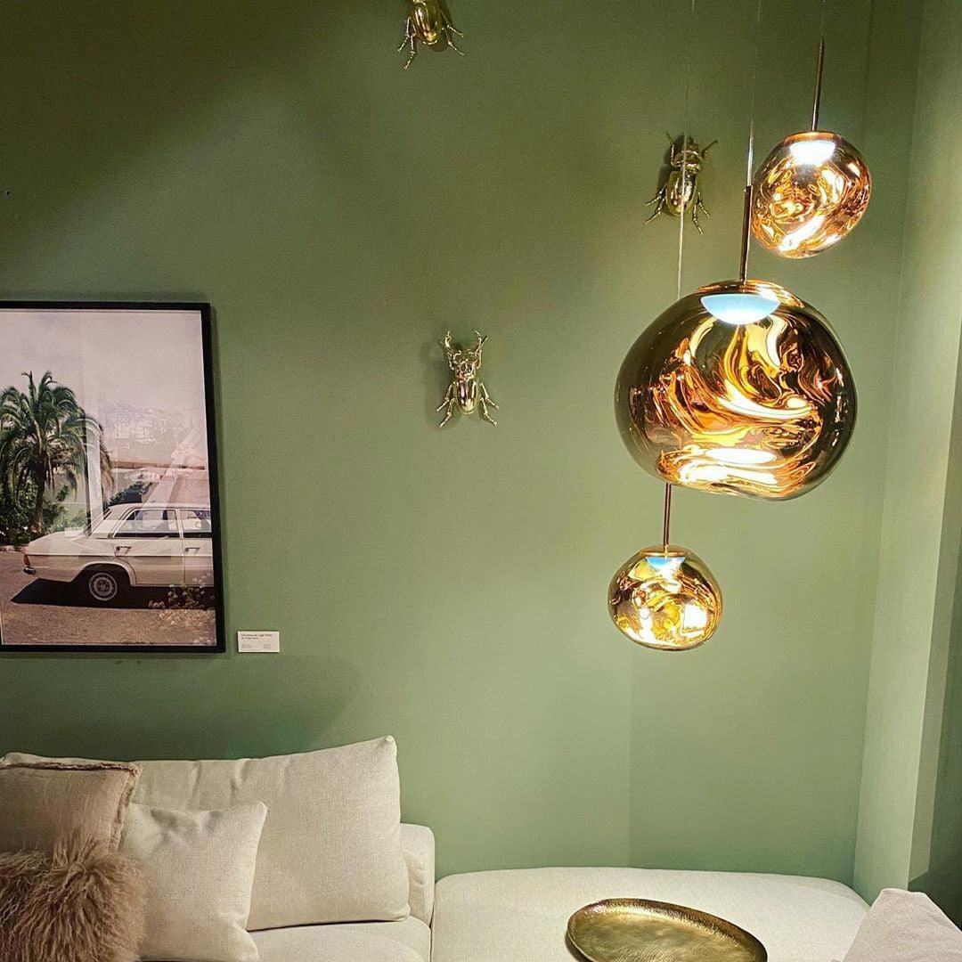 A modern lounge featuring rock pendant lamps with warm reflective gold tones against a sage green wall, adorned with golden insect wall art, a cozy white sofa, and a round metallic coffee table.