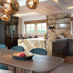 Load image into Gallery viewer, A modern kitchen design featuring rock pendant lamps with warm reflective gold tones, a luxurious marble island with veining, sleek cabinetry, and a dining table with blue chairs and a centerpiece bowl holding fresh fruit.
