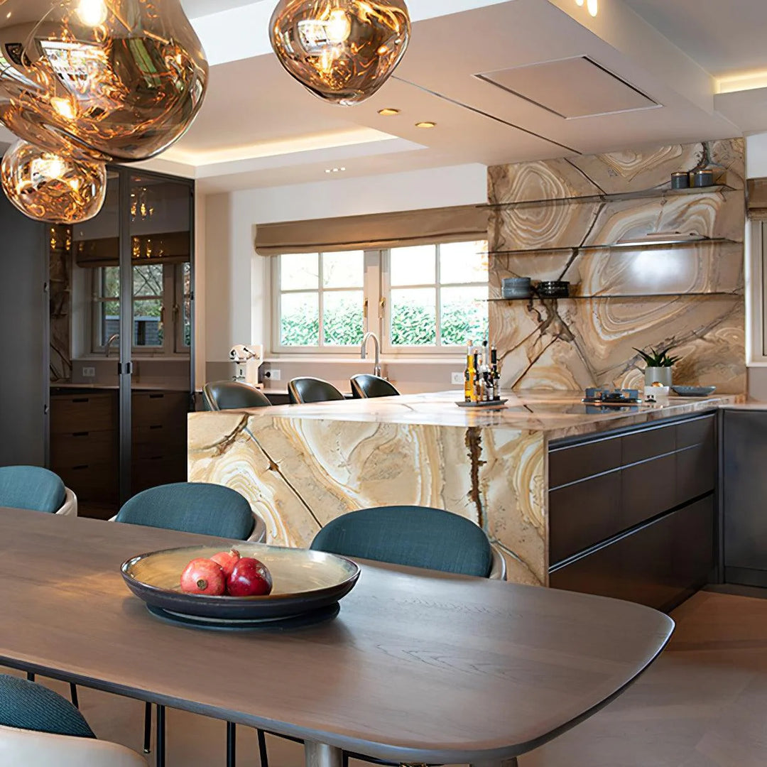 A modern kitchen design featuring rock pendant lamps with warm reflective gold tones, a luxurious marble island with veining, sleek cabinetry, and a dining table with blue chairs and a centerpiece bowl holding fresh fruit.