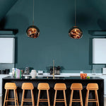 Load image into Gallery viewer, A modern kitchen with two rock pendant lamps featuring reflective gold finishes hanging above a sleek black island countertop, paired with six wooden bar stools and a bold teal accent wall in the background.
