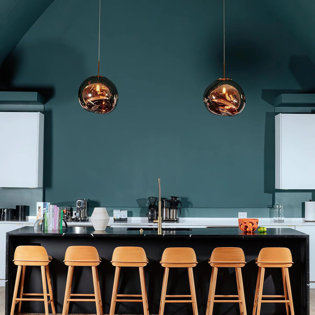 A modern kitchen with two rock pendant lamps featuring reflective gold finishes hanging above a sleek black island countertop, paired with six wooden bar stools and a bold teal accent wall in the background.