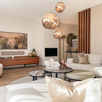 Load image into Gallery viewer, A modern living room featuring rock pendant lamps with warm reflective tones, a neutral curved sofa, layered marble coffee tables, a wooden media console with decorative accents, and a mix of textured cushions and soft lighting.
