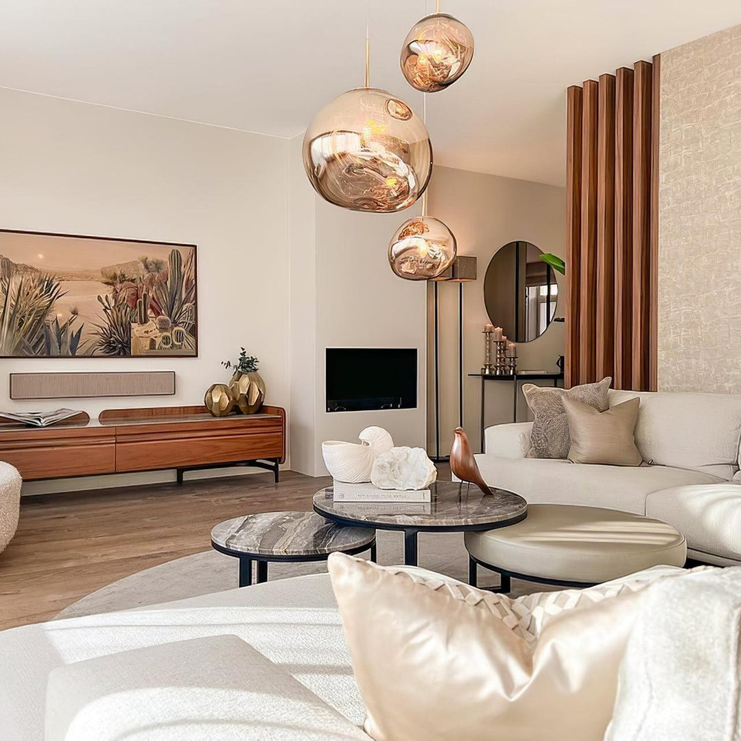 A modern living room featuring rock pendant lamps with warm reflective tones, a neutral curved sofa, layered marble coffee tables, a wooden media console with decorative accents, and a mix of textured cushions and soft lighting.