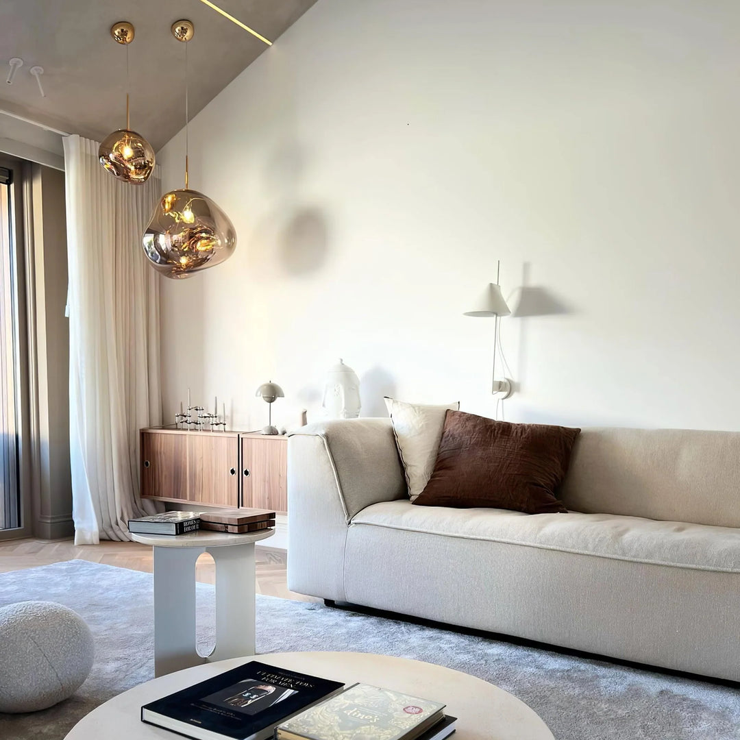 A modern living space featuring rock pendant lamps with reflective gold finishes hanging above a neutral-toned sofa, a minimalist coffee table with books, and a wooden sideboard adorned with decorative objects, complemented by sheer curtains and soft lighting.