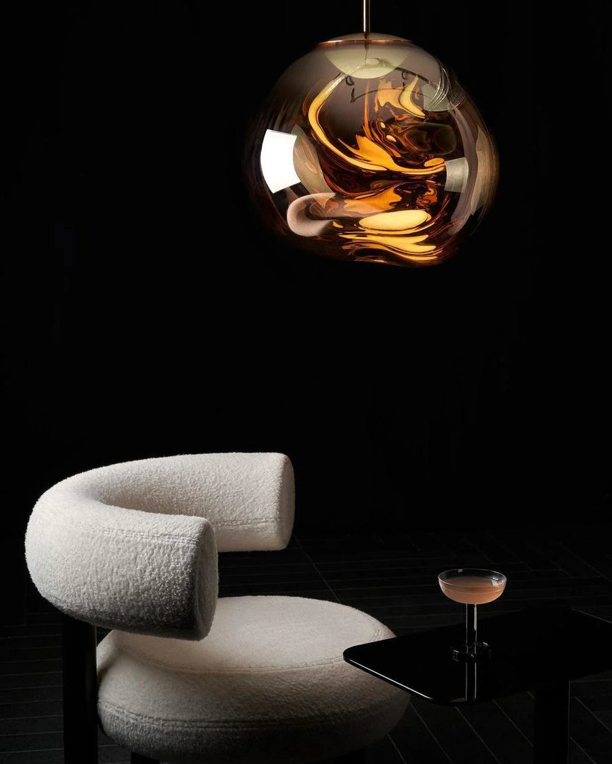 A modern lounge setup with a rock pendant lamp emitting warm reflections above a boucle upholstered chair and a sleek black side table holding a cocktail glass, all set against a dark minimalist background.