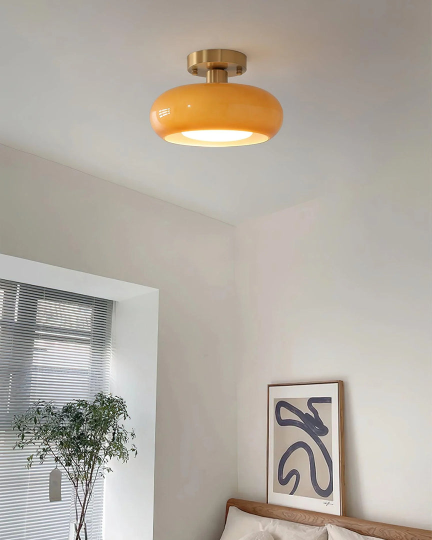 A modern bedroom illuminated by the Sanna cylinder ceiling light with a warm amber shade and brass fixture, complemented by a minimalist wooden bed, abstract wall art, and a lush potted plant near a large window.