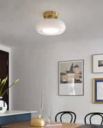 Load image into Gallery viewer, A modern dining area featuring the Sanna cylinder ceiling light with a white shade and brass mount, complemented by a minimalist table setup with decorative accents, black dining chairs, and framed artwork on a neutral wall.
