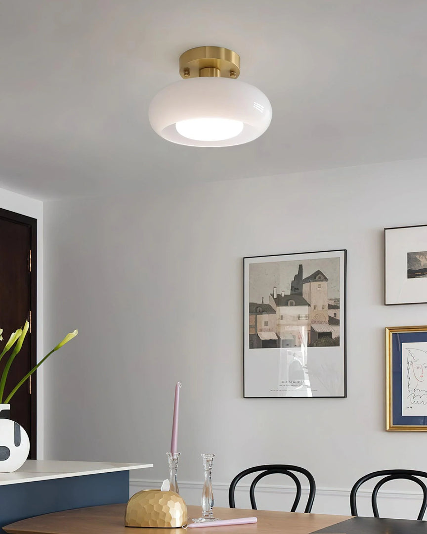 A modern dining area featuring the Sanna cylinder ceiling light with a white shade and brass mount, complemented by a minimalist table setup with decorative accents, black dining chairs, and framed artwork on a neutral wall.