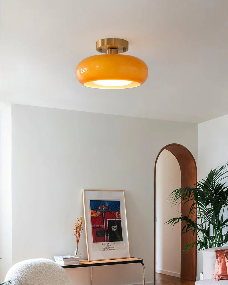 A modern living space featuring the Sanna cylinder ceiling light with a warm amber shade and brass mount, complemented by a sleek console table with decorative items, an arched doorway, and lush green plants adding a vibrant touch.