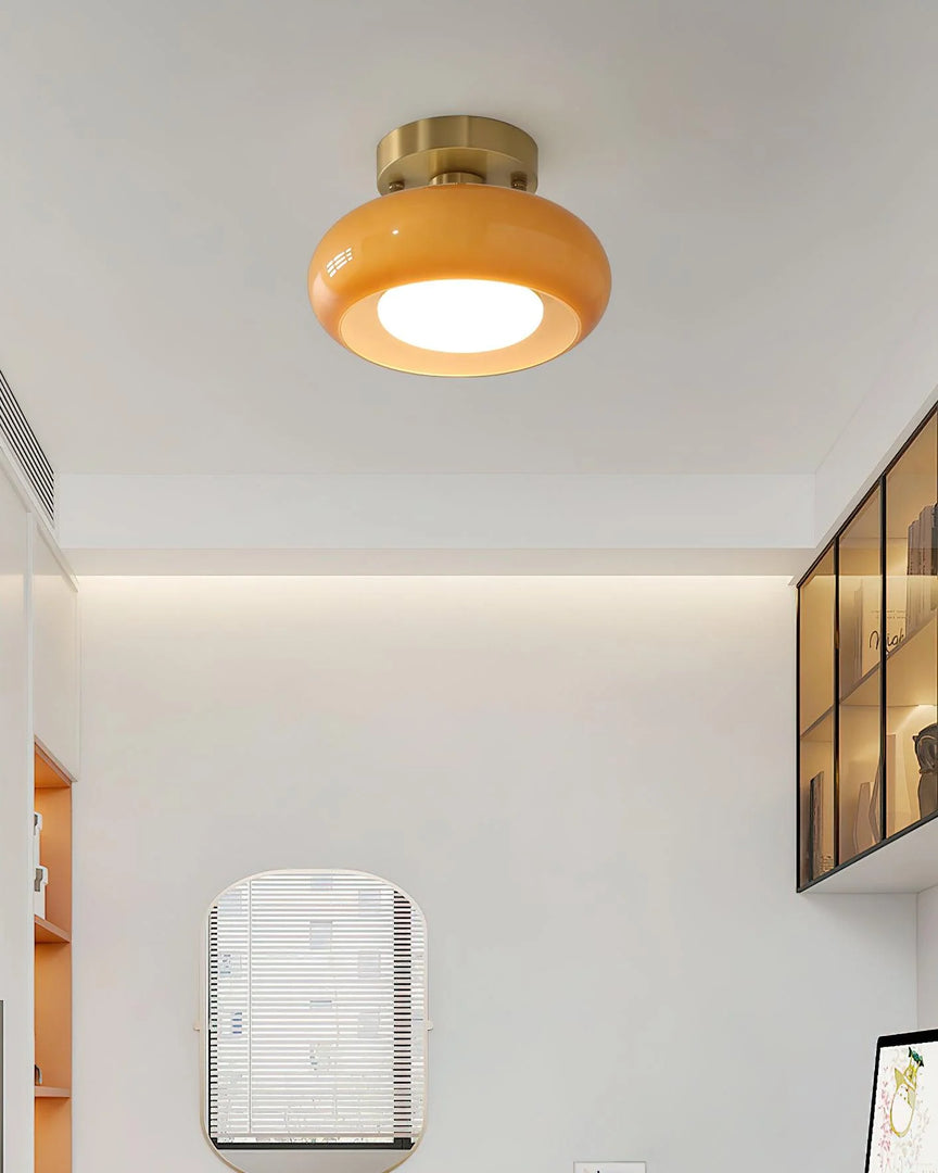 A contemporary study room illuminated by the Sanna cylinder ceiling light with a warm amber shade and brass mount, featuring a minimalist design with built-in shelving, a desk, and an arched mirror on a clean white wall.