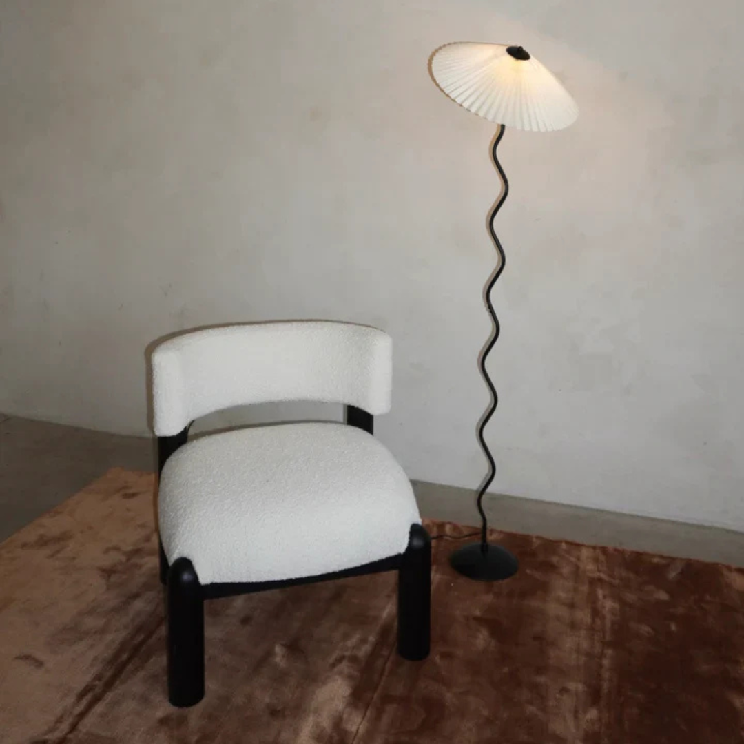 A minimalist reading corner featuring the Seashell floor lamp with a sculptural wavy stand and pleated shade, paired with a modern boucle accent chair on a soft brown rug against a neutral wall.