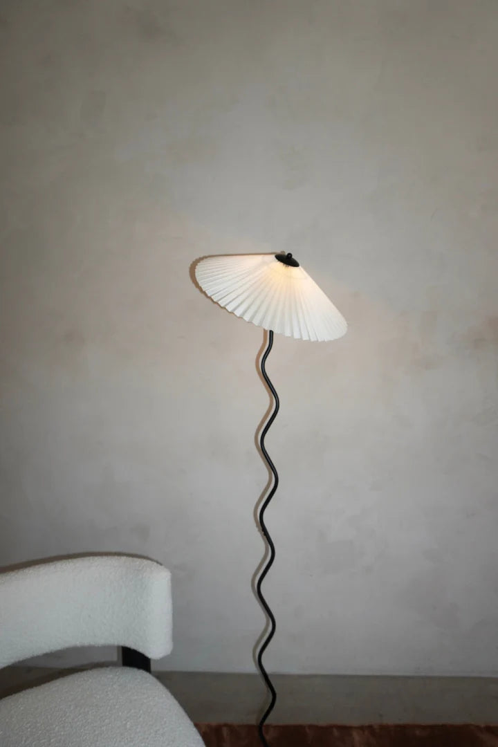 The Seashell floor lamp with a pleated shade and wavy stand placed next to a boucle accent chair on a warm brown rug, set against a minimalist neutral wall for a modern and cozy aesthetic.